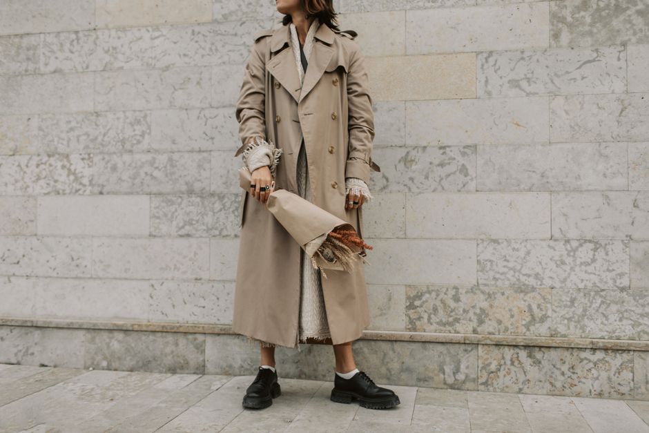 Stylish woman in beige trench coat holding a bouquet against a marble wall. Elegant urban fashion vibe.