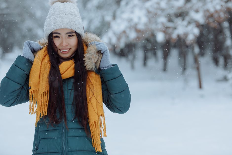 Asian woman smiling in snowy winter with bright scarf and beanie in an outdoor setting.