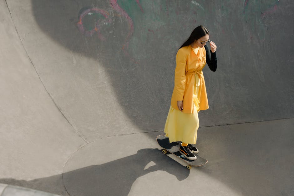 Female skater in a vibrant yellow dress rides a skateboard in a sunny outdoor skate park.