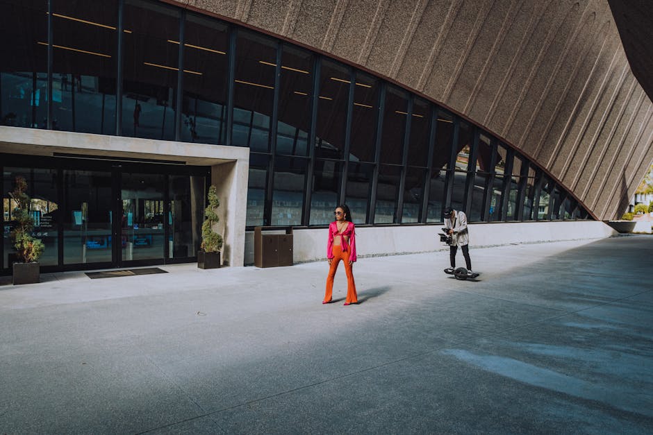 A fashionable photoshoot in front of a modern architectural facade featuring a model and a videographer.