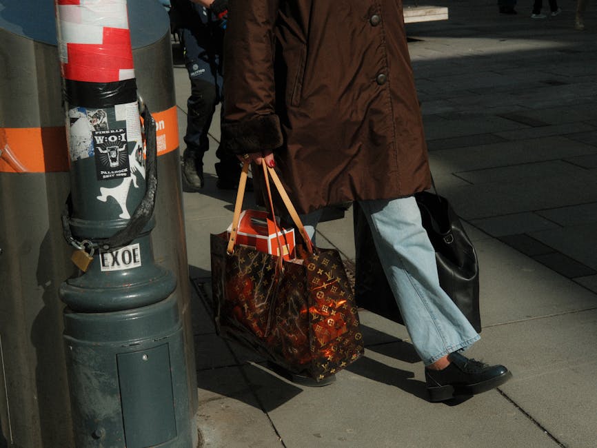 A person in stylish attire walks with luxury brand shopping bags on a city street.