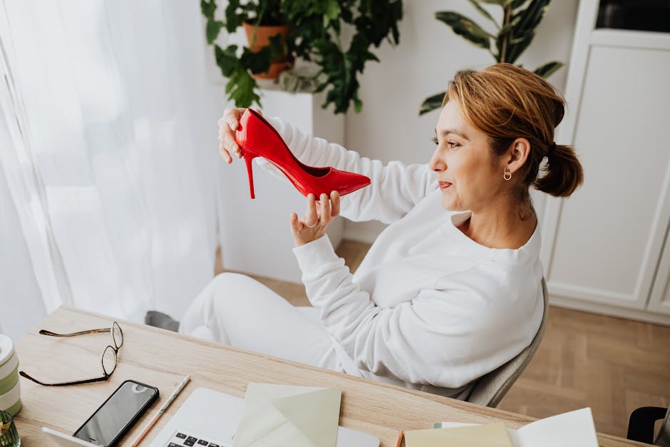 Woman in white sweater smiling at red high heel shoe at a desk with plants.