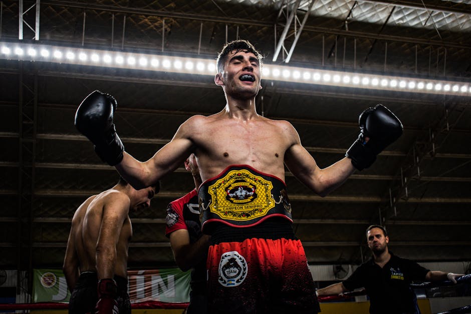 A triumphant boxer with championship belt raises his arms in victory inside a boxing ring.