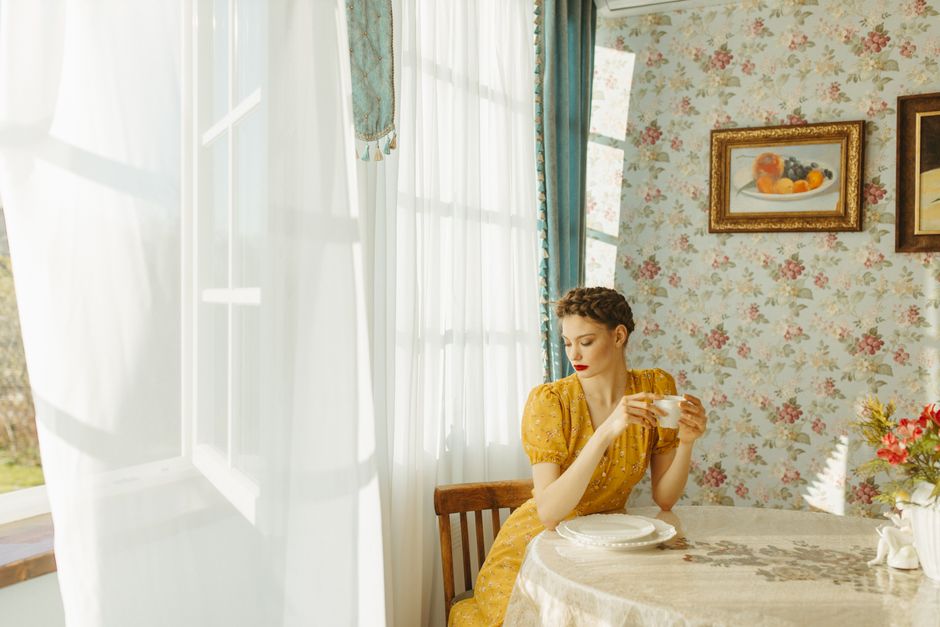 A woman in a yellow dress enjoys coffee by a sunlit window in a vintage room.