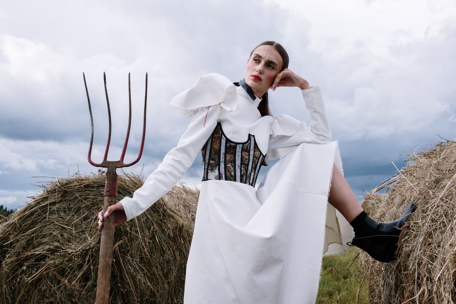 Stylish woman in white dress posing with a pitchfork beside hay bales, exuding modern rural fashion.
