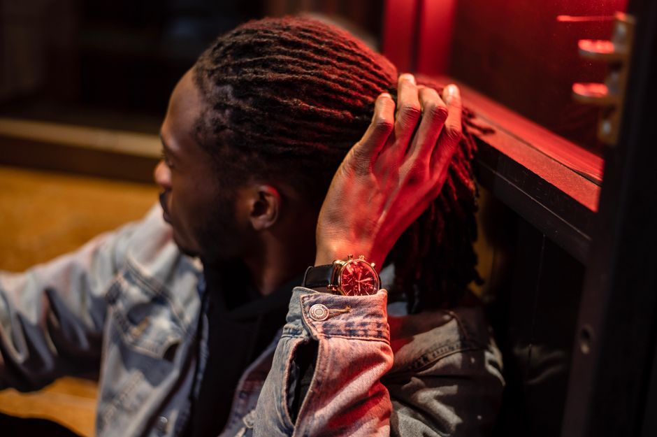 High angle of serious African American male touching dreadlocks while sitting near building with red illumination