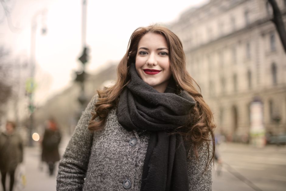 Happy woman in a gray coat and scarf standing outdoors, showcasing a charming smile on a c