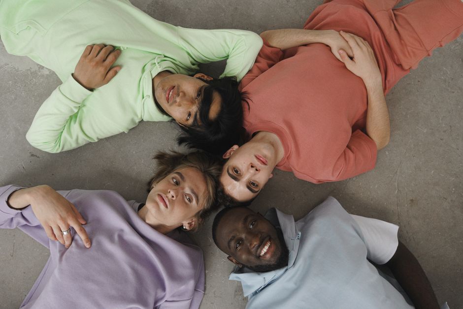 Four young adults lying on the floor in casual pastel outfits, forming a circle.