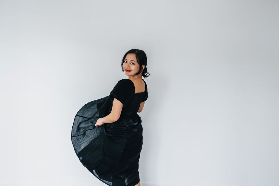 An elegant woman in a black dress poses gracefully indoors against a plain background.
