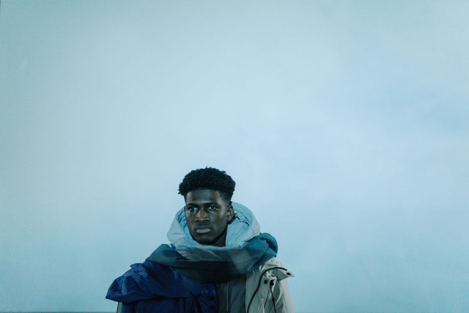 Man in a winter jacket and scarf, posing against a pastel blue studio background.