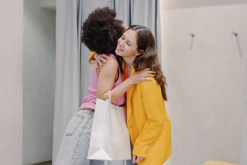 Two young women embrace in a fitting room. One holds a shopping bag, expressing happiness and friendship.