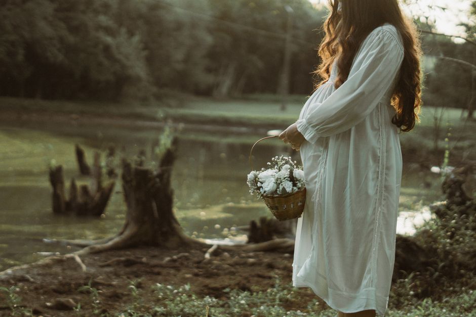 Pregnant woman in a white dress holds a basket of flowers by a peaceful river setting.