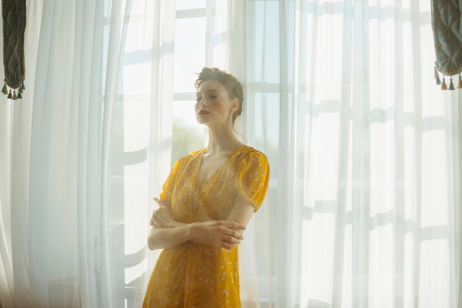 A woman in a yellow dress stands gracefully by sunlit curtains, embodying elegance and serenity.