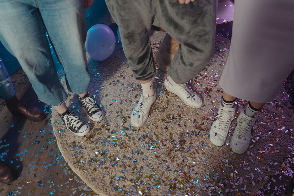 Casual party scene with people wearing sneakers, surrounded by confetti and balloons indoors.
