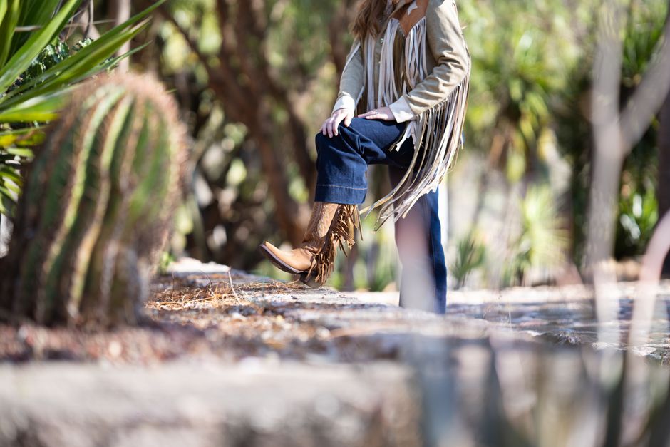 A woman in a fringe jacket and jeans poses near a cactus in a sunny outdoor scene.